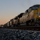 A yellow freight train going down the tracks backlit by a sunset.